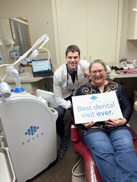 Dentist and patient in dental office, patient holds sign: "Best dental visit ever." Both smile.