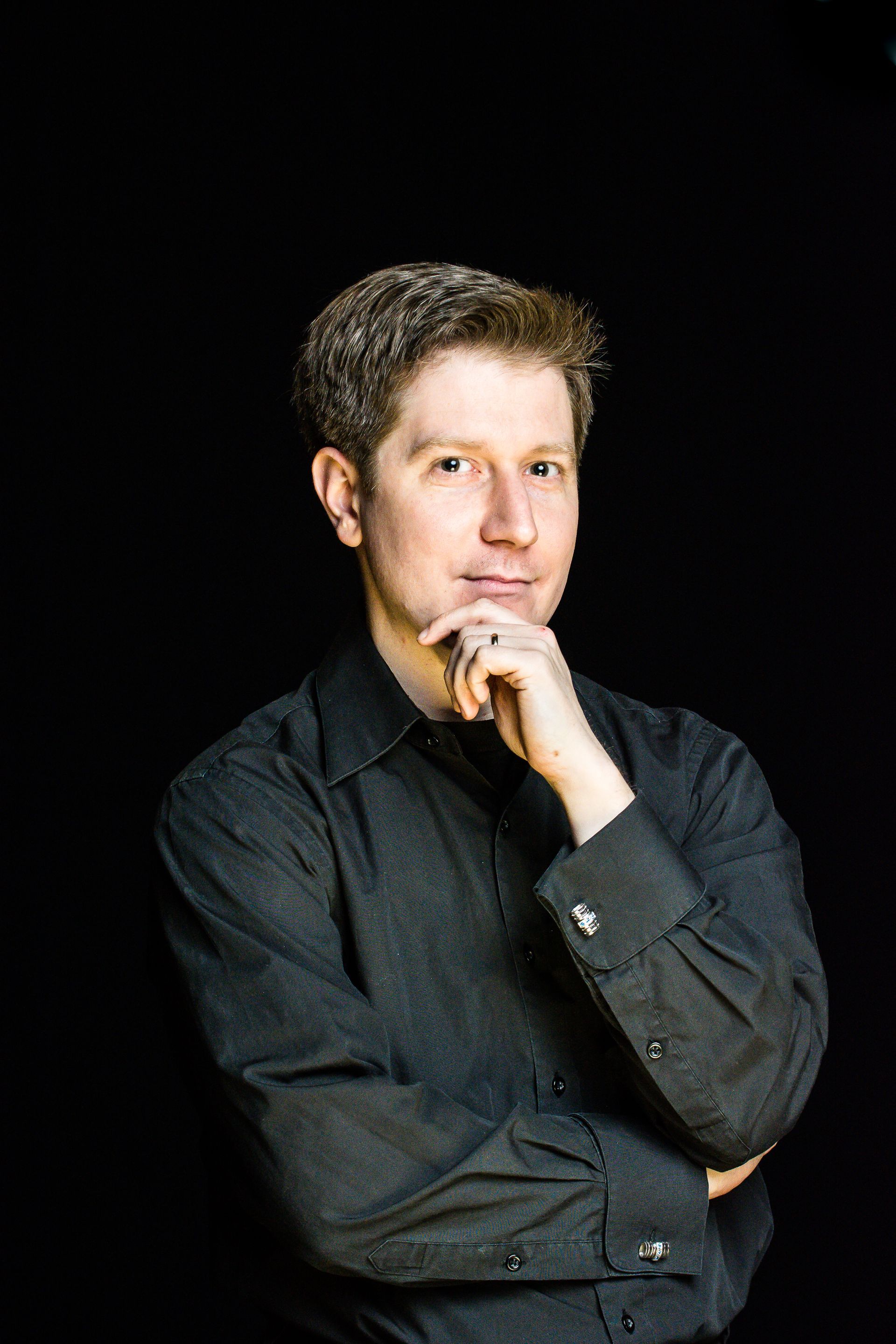 A person in a black button-down shirt rests their chin on their hand against a plain black background.