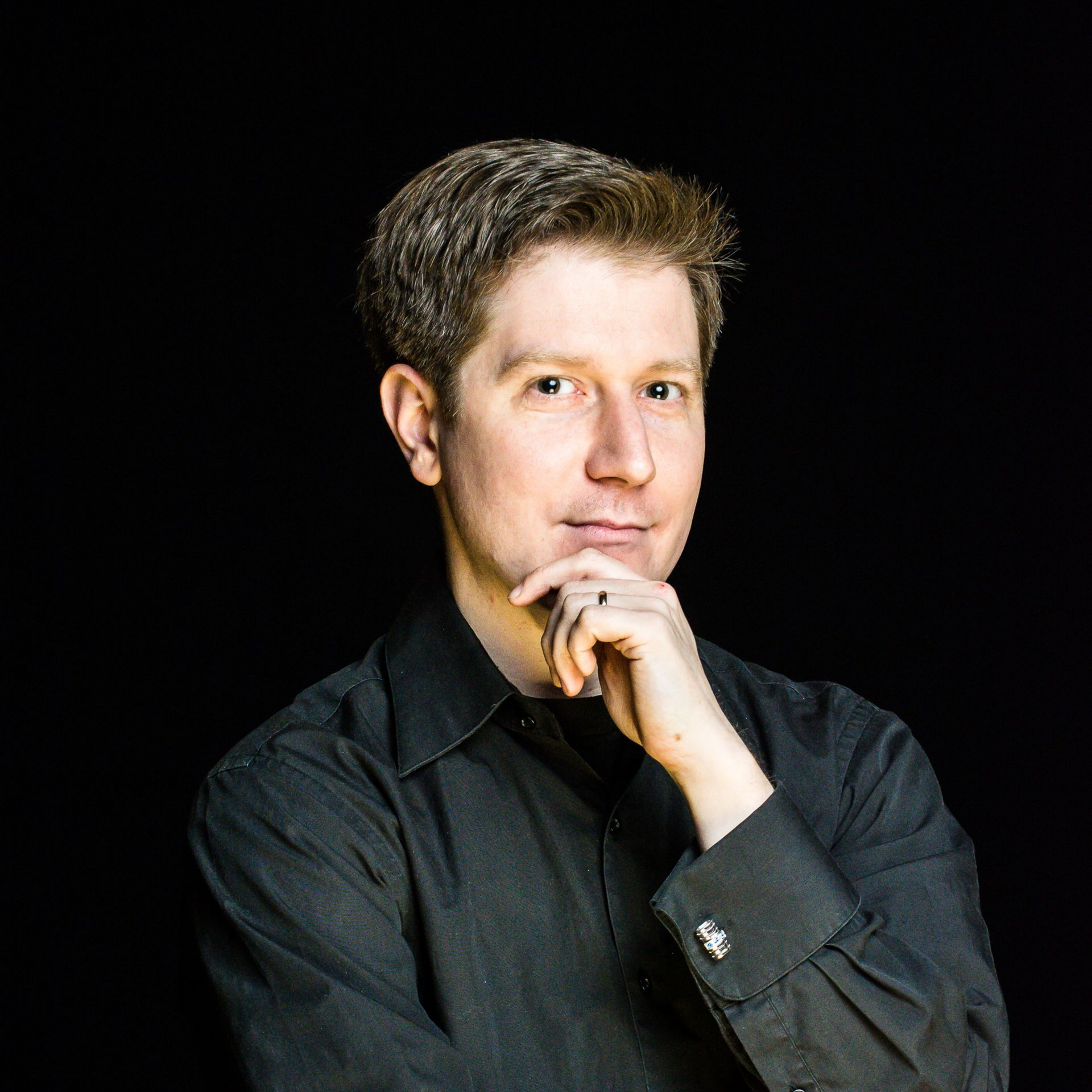 A person in a black button-down shirt poses against a black background, resting their chin on their hand.