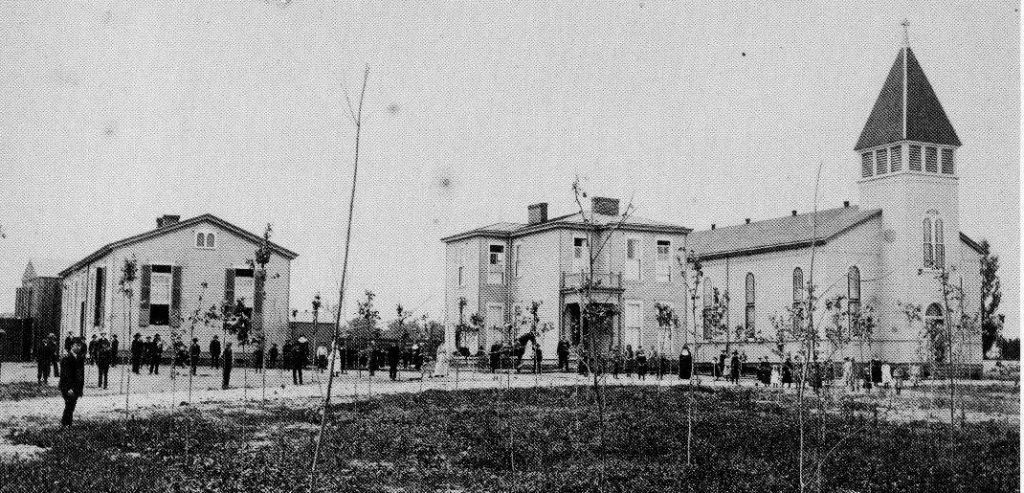 A historic, black-and-white photograph showing two stone buildings, including a church with a tower, and a group of people.