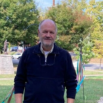 A person with a beard and glasses tucked into a dark quarter-zip sweater stands outdoors in a tree-filled park.