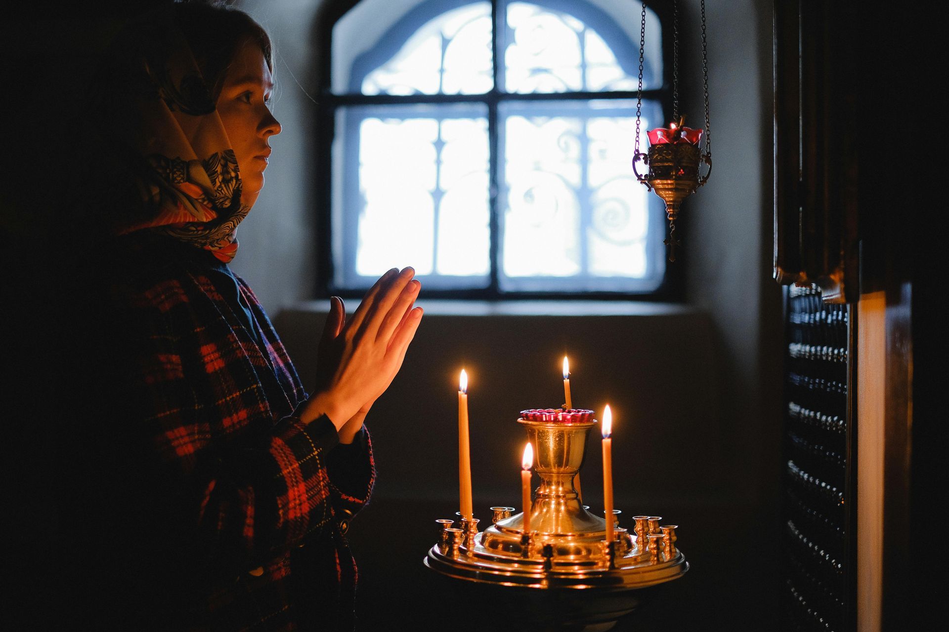 A person stands with hands pressed together in prayer before a metal candle stand with lit candles inside a dim church.
