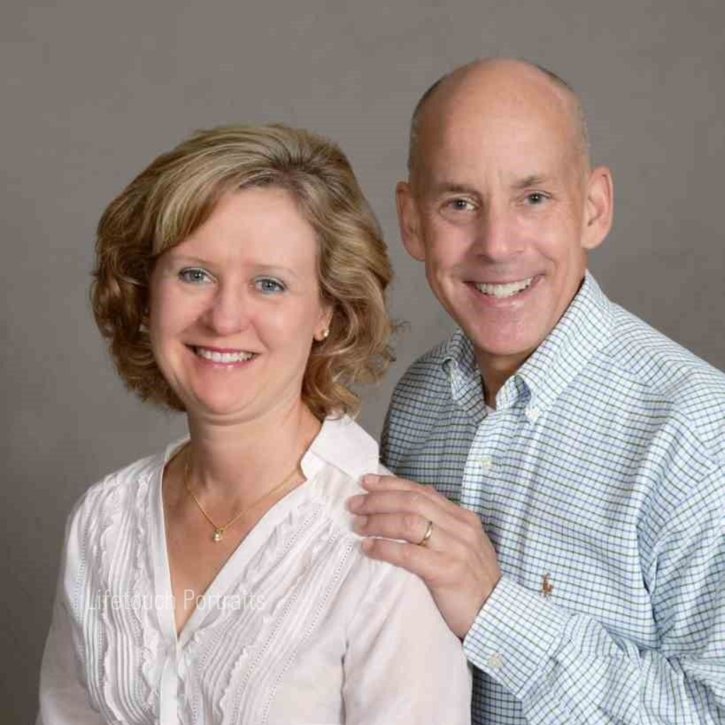 A smiling couple posing for a portrait against a gray backdrop; he has his hand gently on her shoulder.