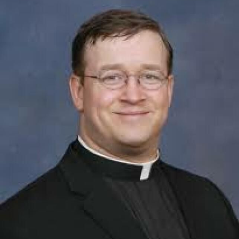 Headshot of a person wearing glasses and a black clerical shirt with a white tab collar, set against a blue background.