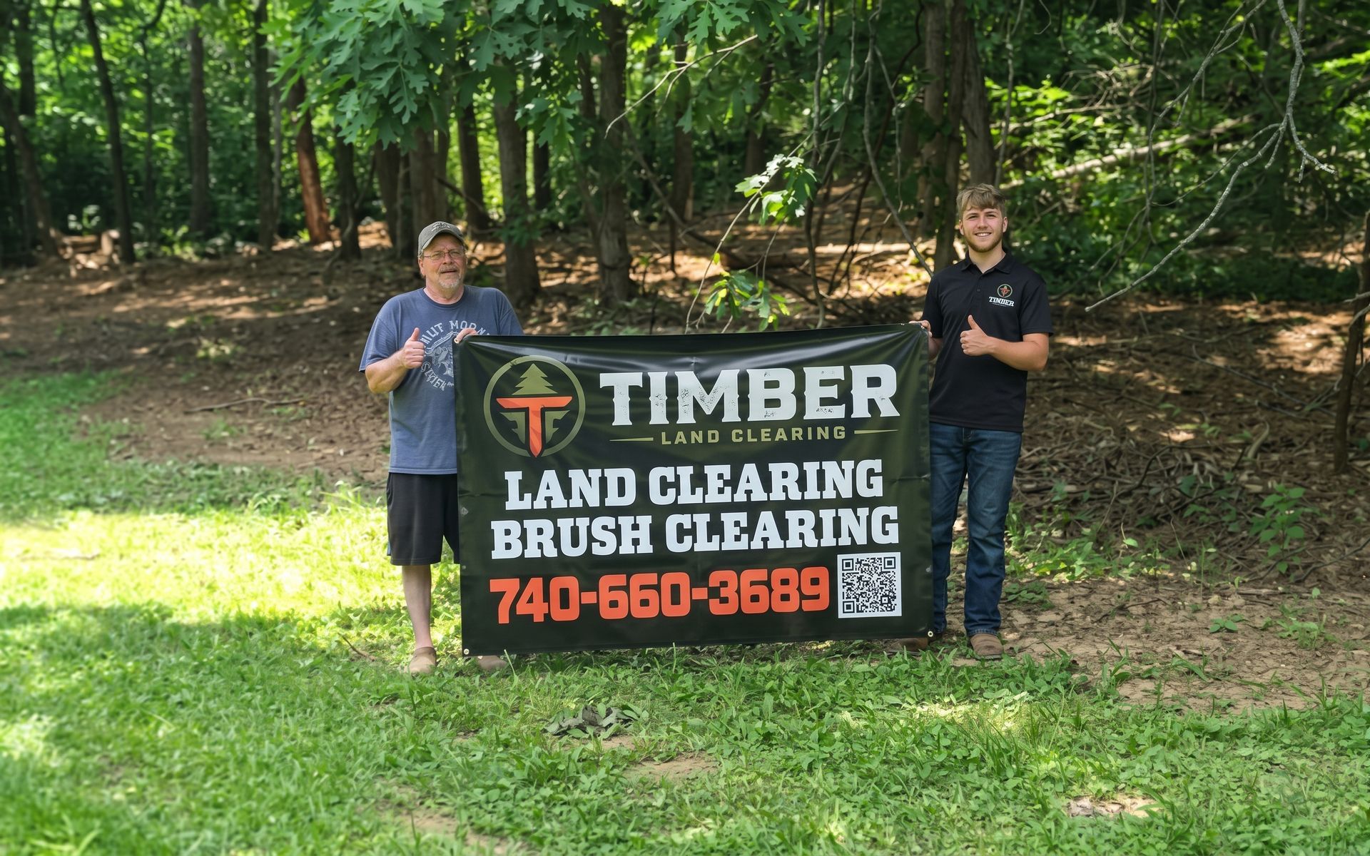 Two people holding a Timber Land Clearing sign in a wooded area with green grass.