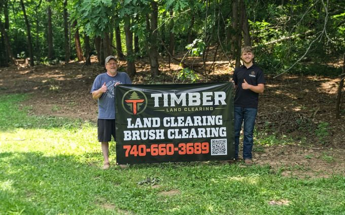 Two people holding a Timber Land Clearing sign in a wooded area with green grass.