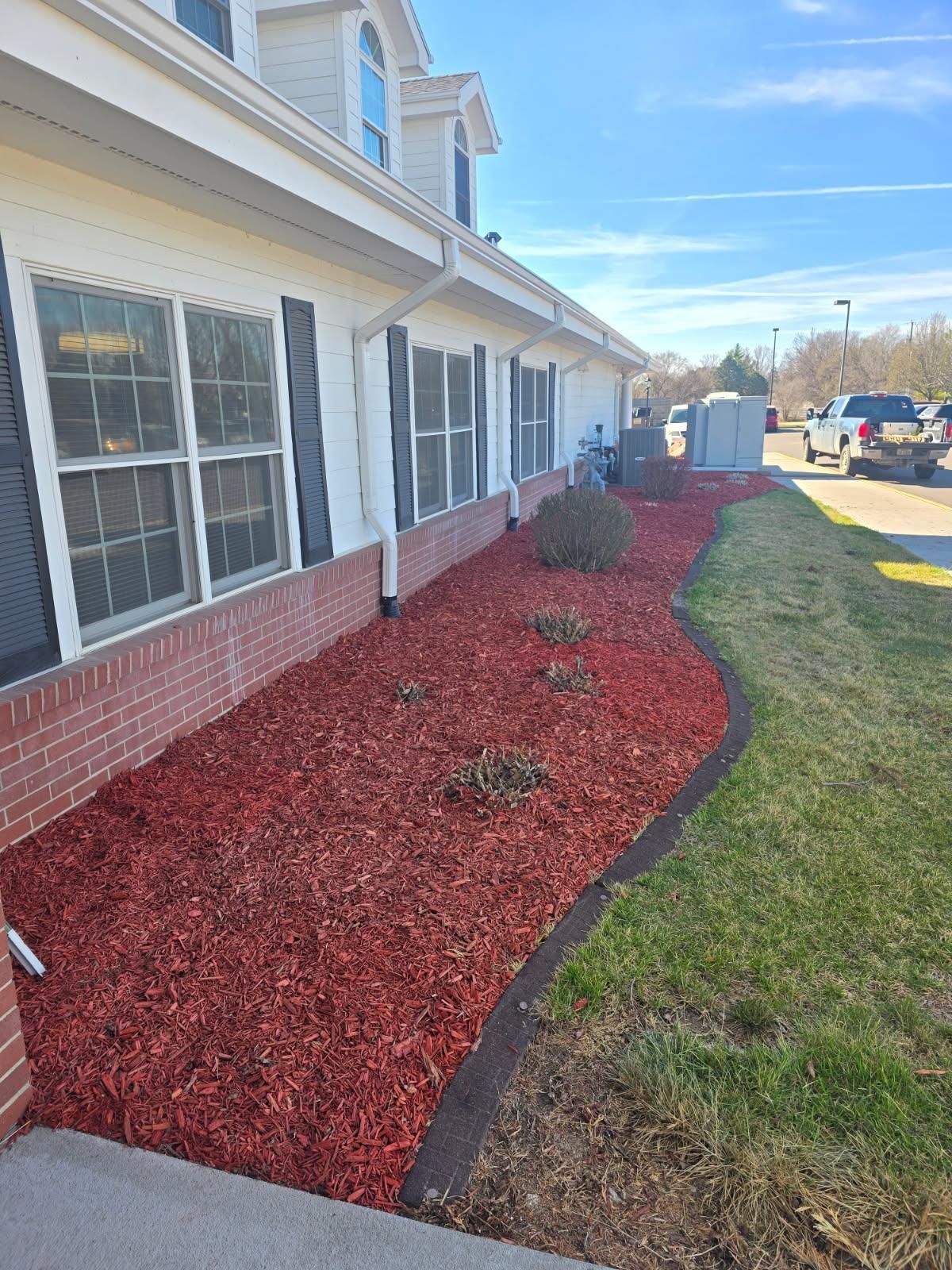 A tan building with a brick base and red mulch garden bed featuring small shrubs along the side.