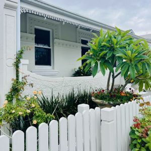 A white picket fence surrounds a garden in front of a white house.