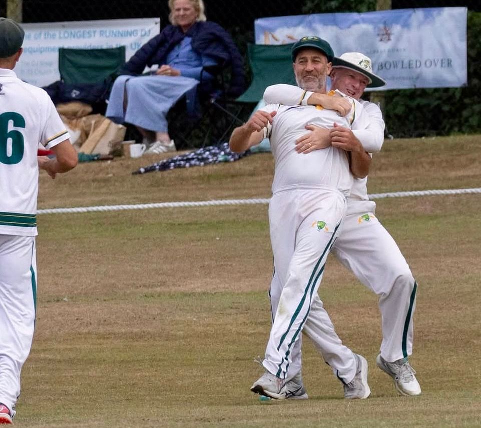 Two cricket players in white uniforms embrace on the field, celebrating