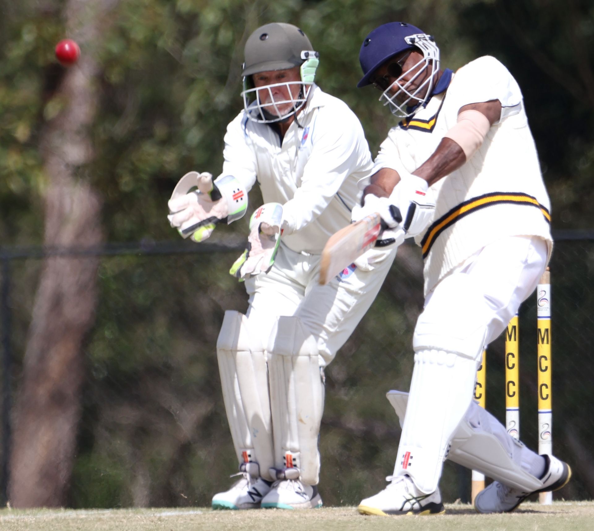 Cricketer batting, hitting a red ball. Wicketkeeper crouched, ready to catch. Sunny outdoor field.