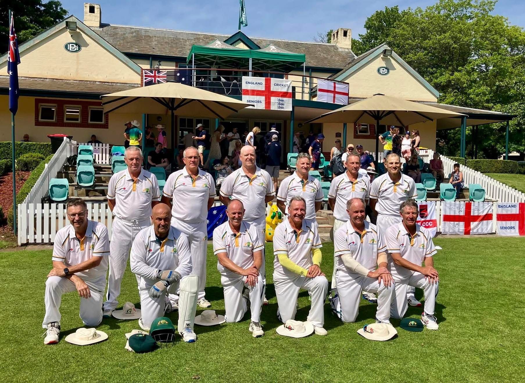 Cricket team in white with gold trim, kneeling in front of a clubhouse
