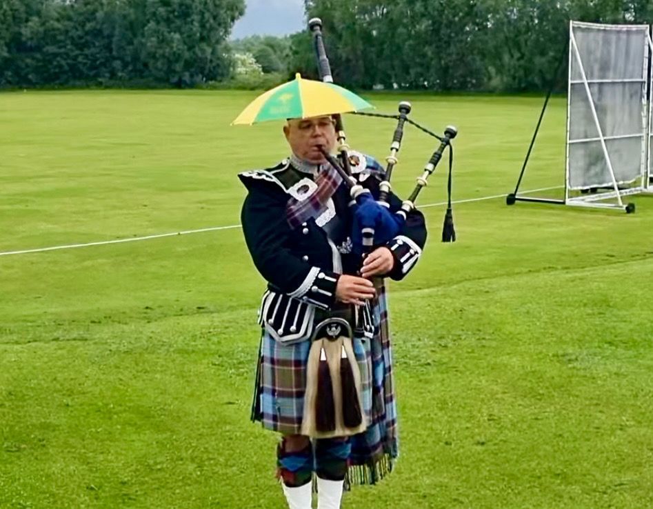 Bagpiper in traditional attire, playing on a field with a yellow and green umbrella on the head