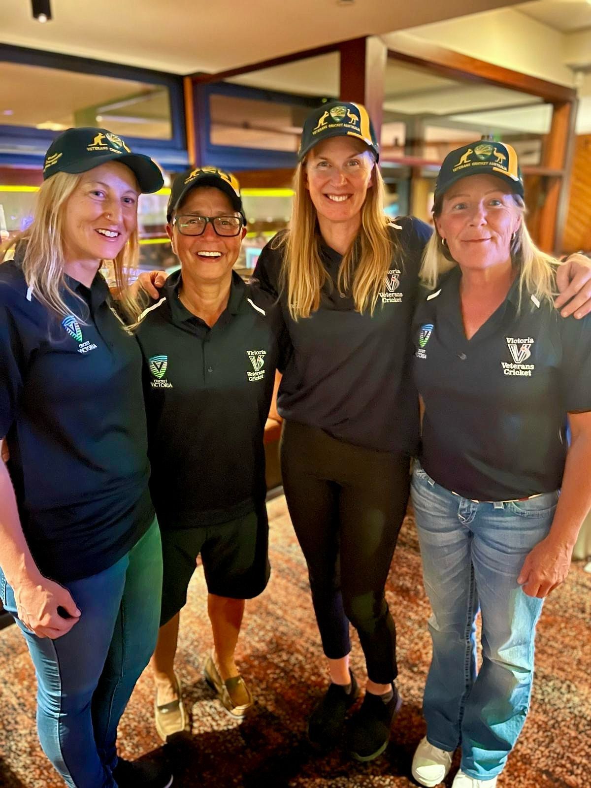 Four people, wearing matching caps and shirts, standing indoors, smiling, arms around each other.