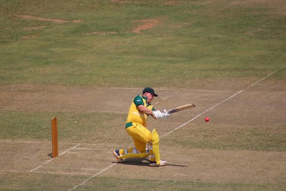 Cricketer in yellow and green uniform batting at a cricket pitch, hitting a red ball