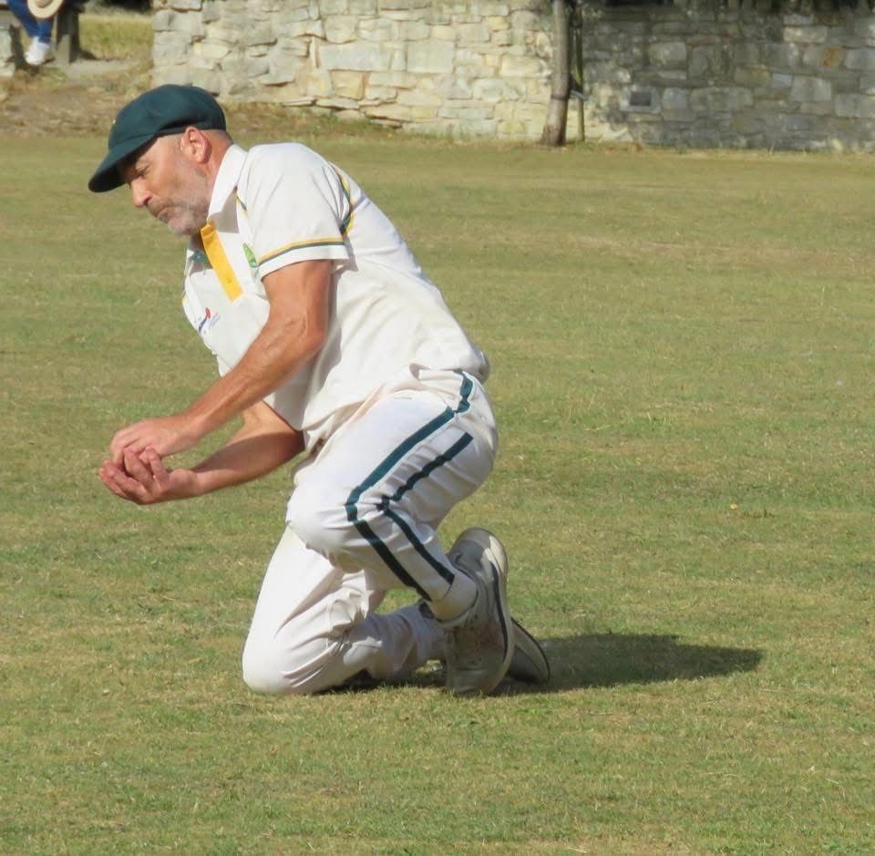 Man in cricket whites kneels on grass, catching a ball