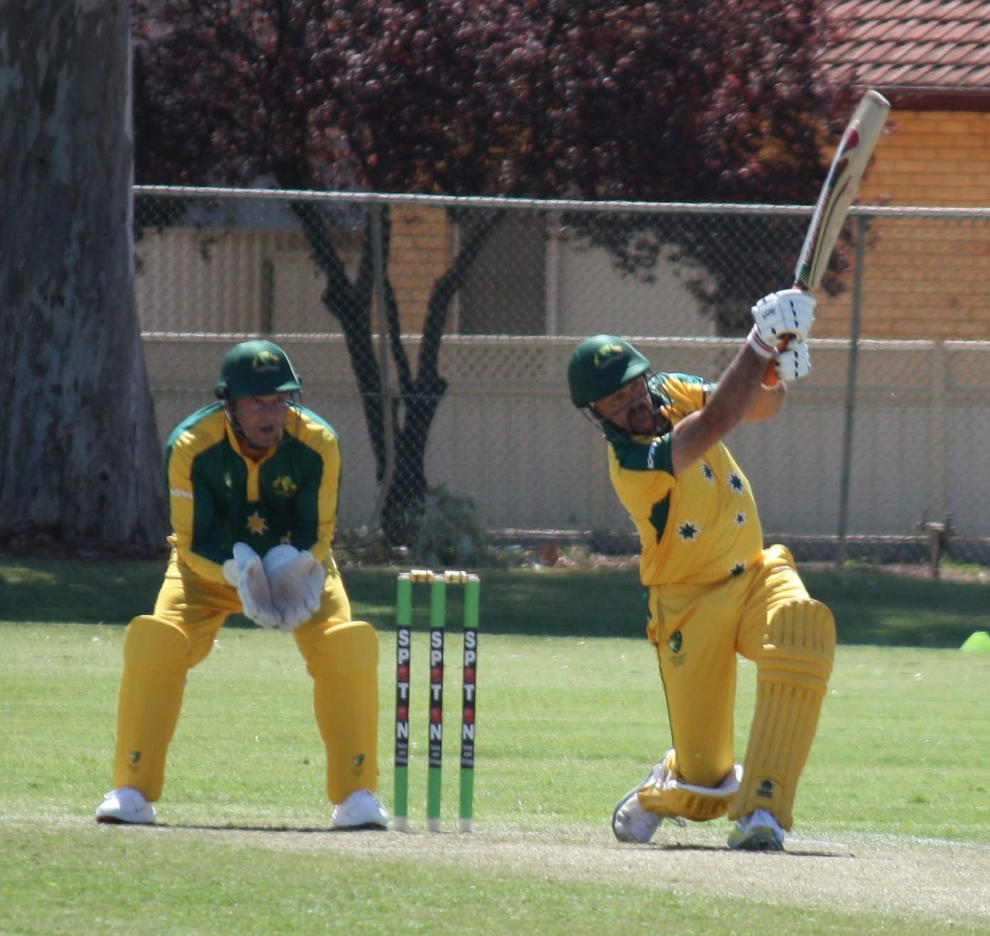 Cricket batsman in yellow uniform hitting a ball, wicketkeeper behind the wickets