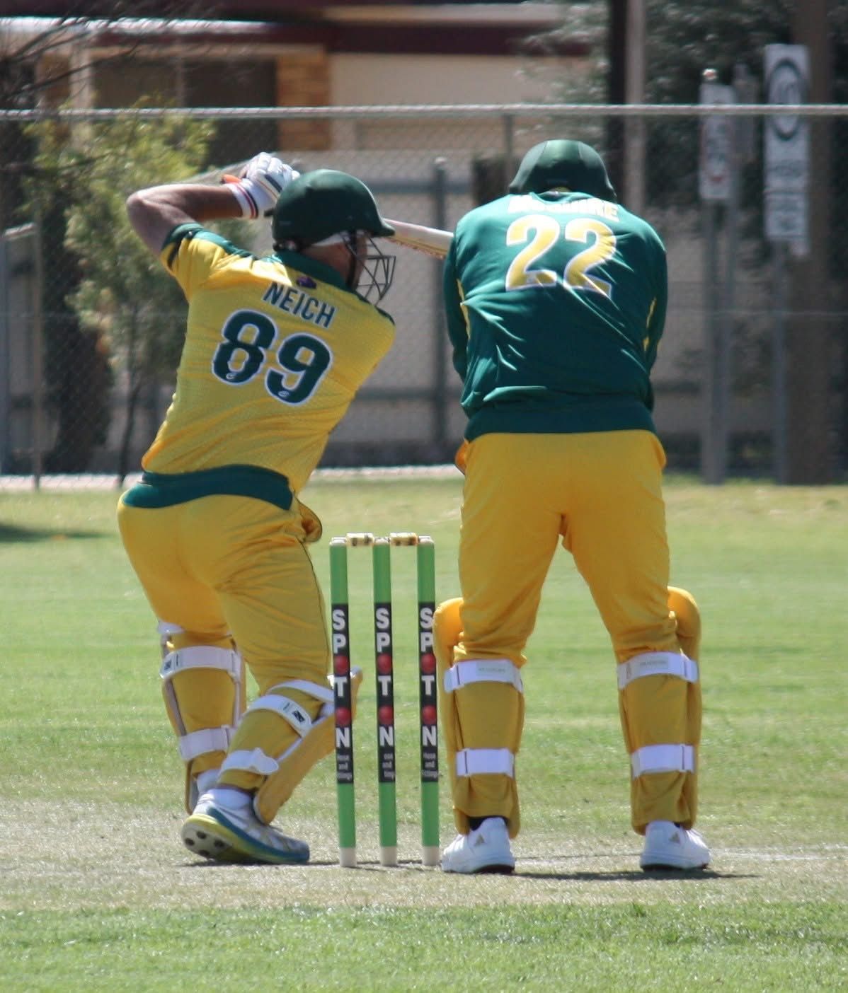 Cricketer in yellow and green uniform batting. Wicket keeper behind