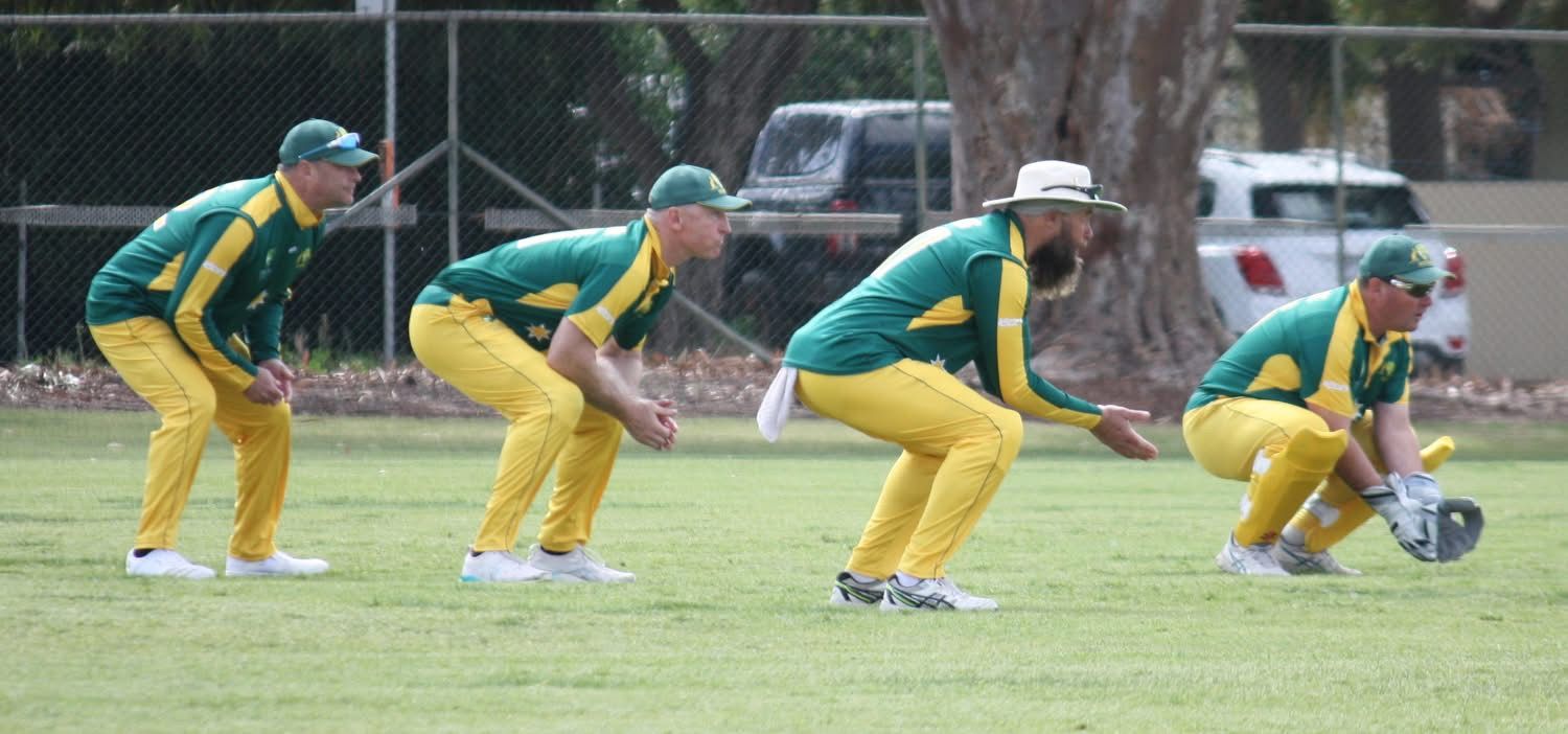 Cricket players in green and yellow uniforms squat on a grassy field