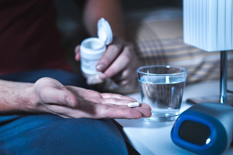 Person holding pill and pouring from a pill bottle, with a glass of water and a clock on a nightstand.