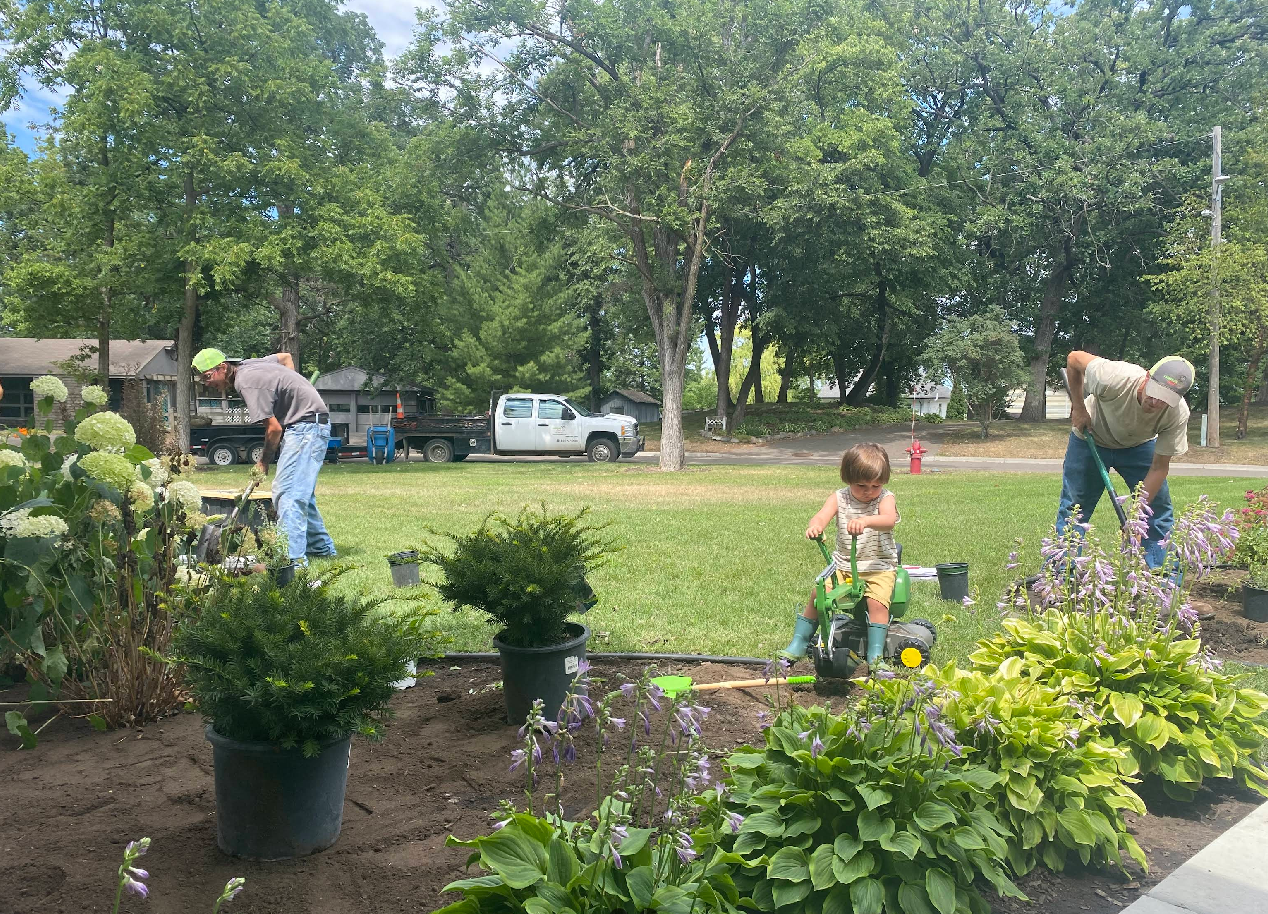 Three adults and a child gardening in a yard on a sunny day. The child pushes a toy lawnmower.