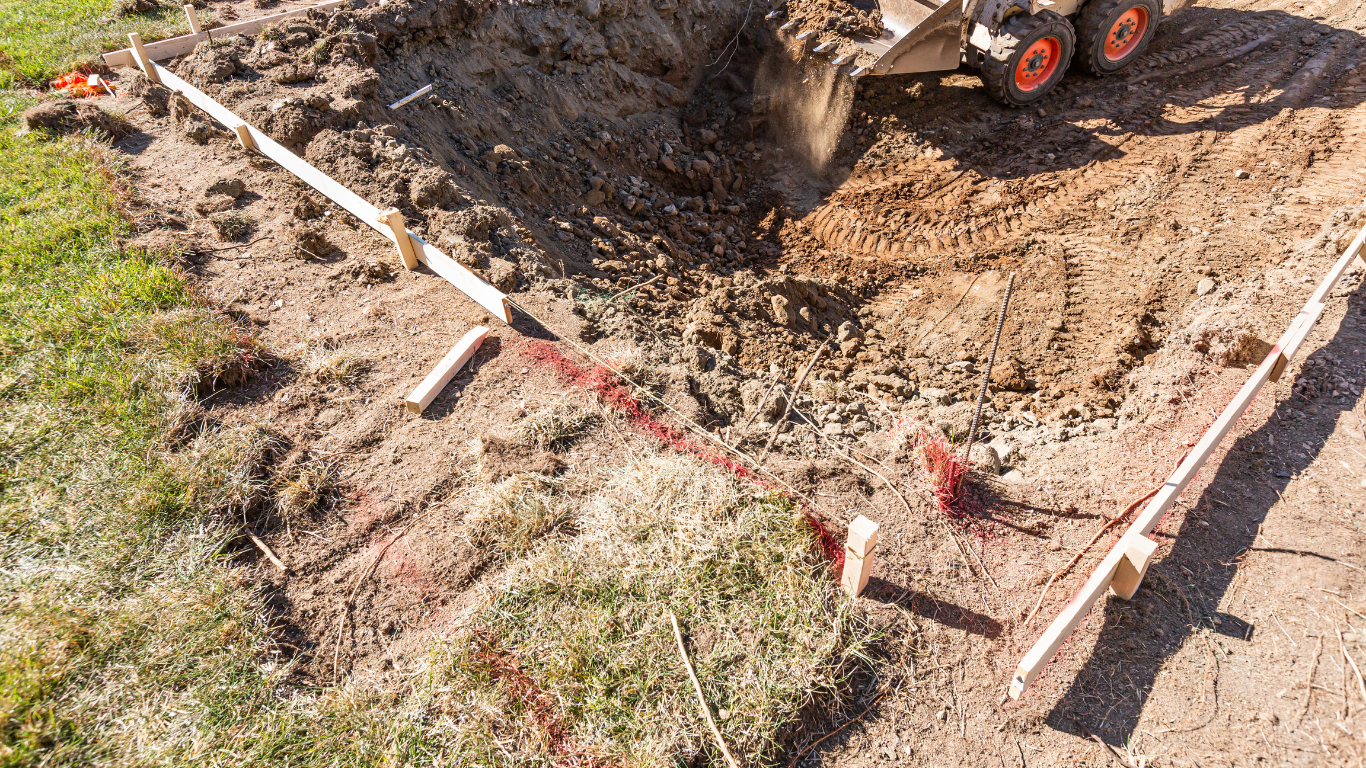 A small excavator digging a hole within a square-marked area outlined by wooden stakes and string on a dirt surface.