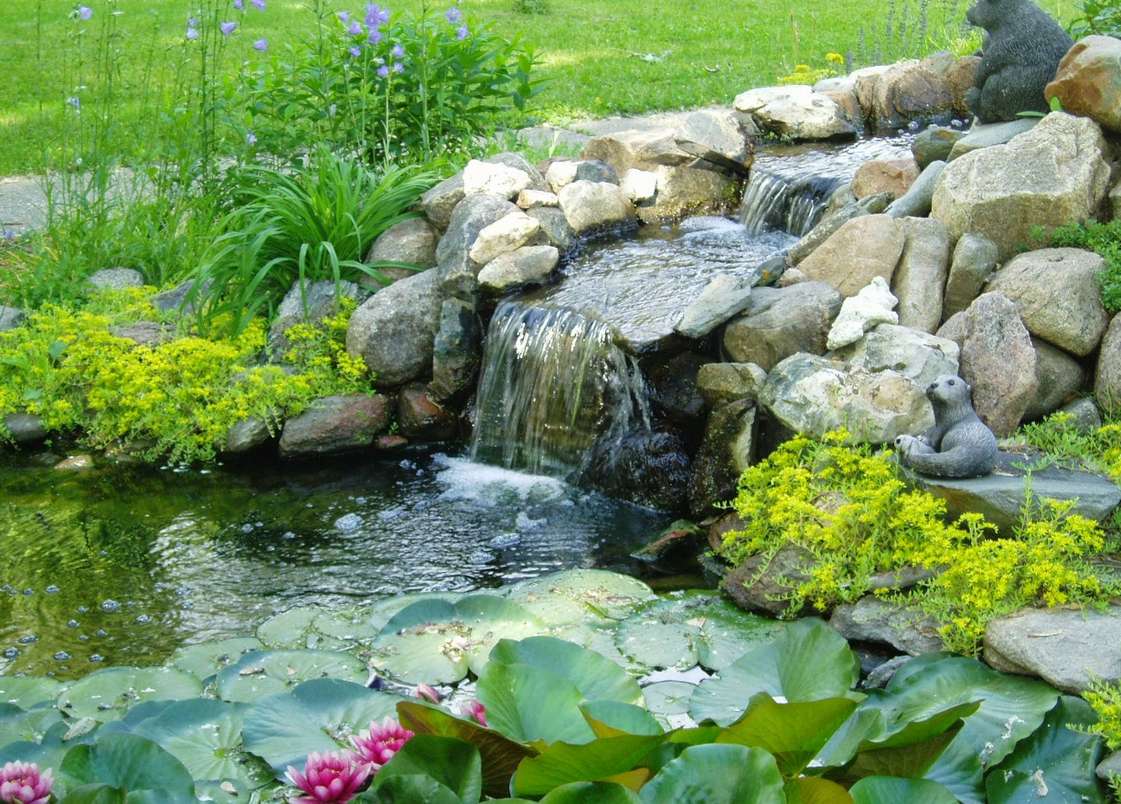 Water cascades over rocks into a pond with lily pads and a small sculpture.
