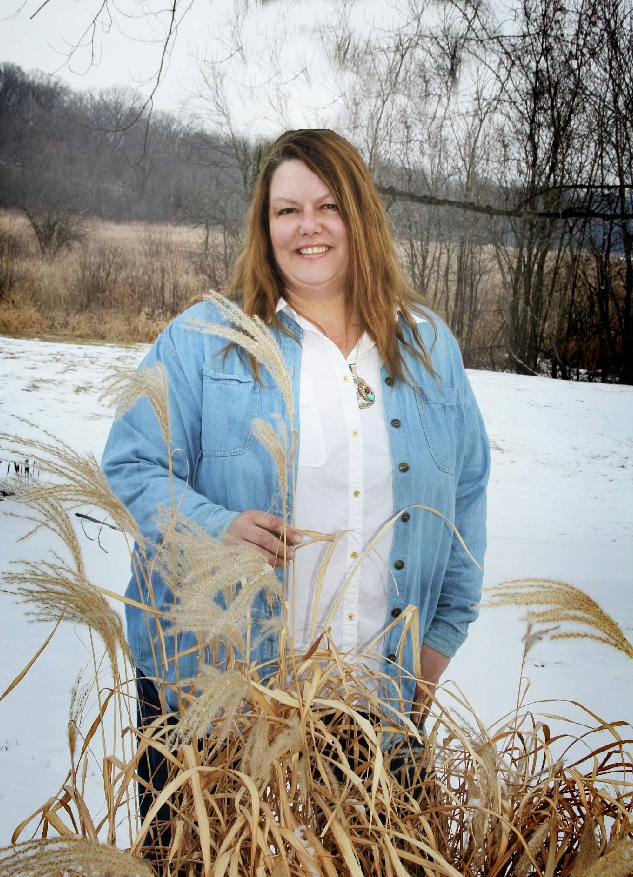 Woman in denim shirt and white shirt poses in snowy field, holding tall dried grasses.