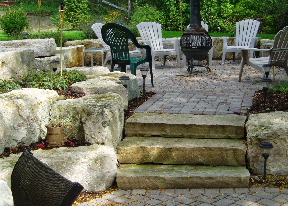 Stone steps leading to a patio with seating, a fire pit, and large rock landscaping.