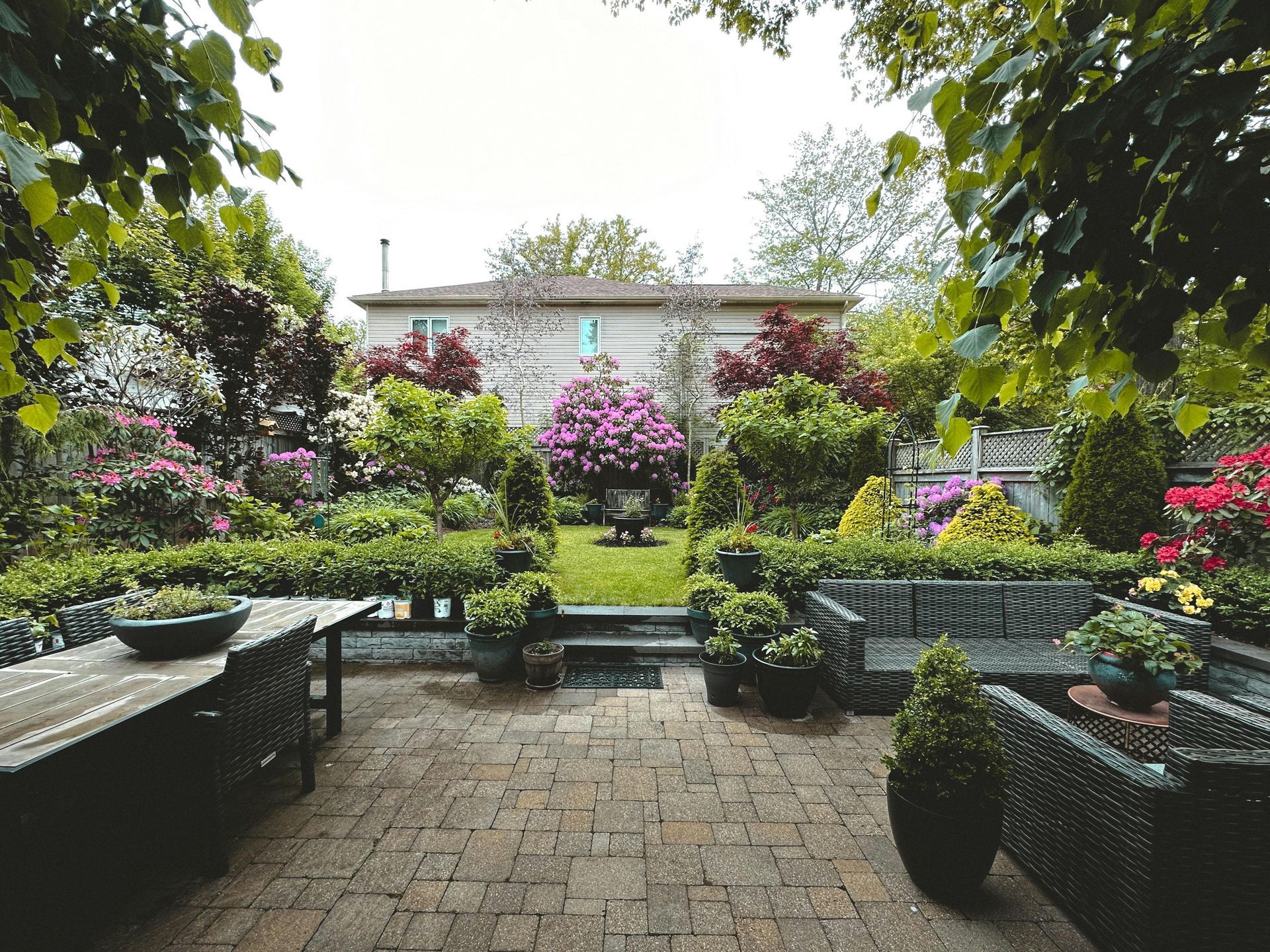 A patio with stone pavers, wicker furniture, and potted plants overlooking a lush backyard garden with a house beyond.