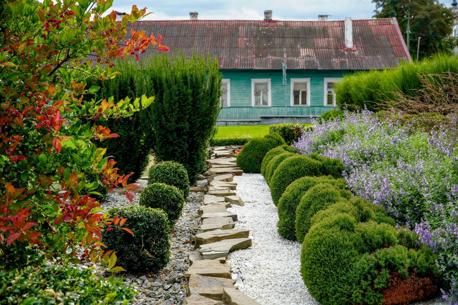 A stone path cuts through a garden of manicured shrubs and white gravel, leading toward a teal wooden house.