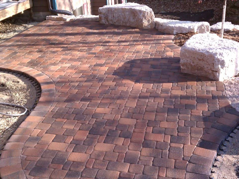 Brick patio with reddish-brown pavers, curved edges, and large light-colored boulders.