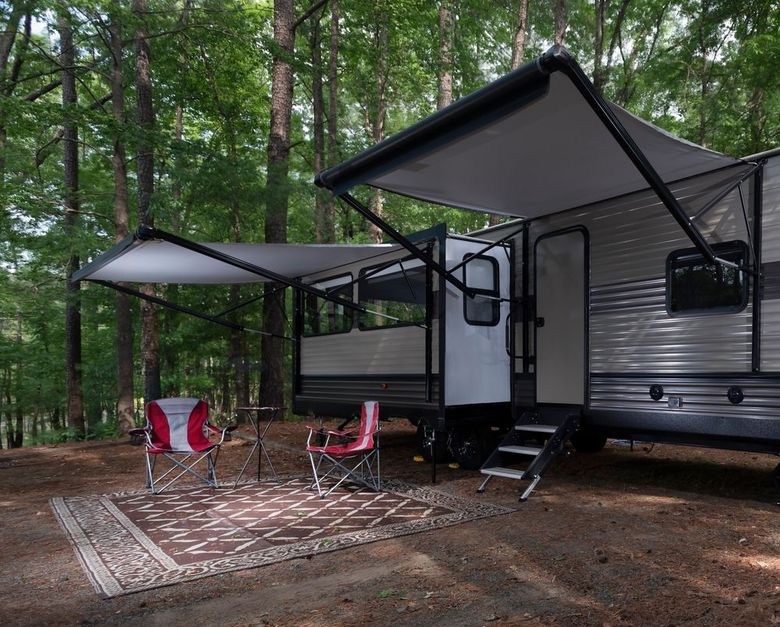 Covered RV campsite with awning, patio rug, and two red chairs in a wooded area.