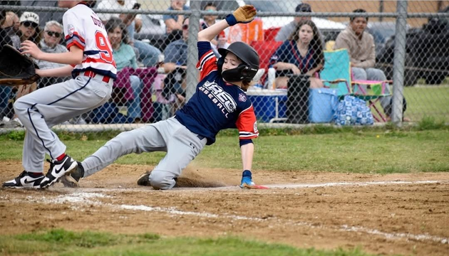 Baseball player sliding into home plate, catcher in front. Blue and gold uniforms, action shot.