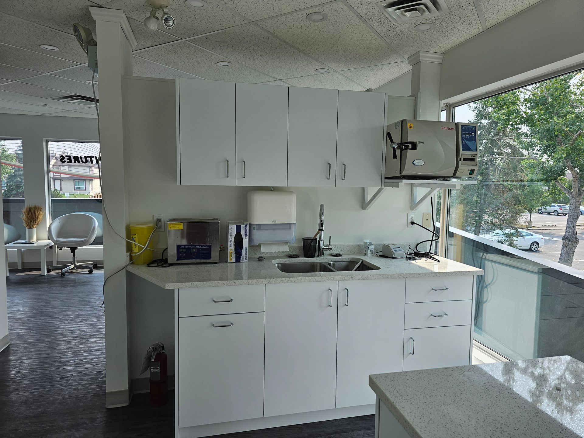 Small white kitchen with cabinets, sink, microwave, and a view of trees through large windows.