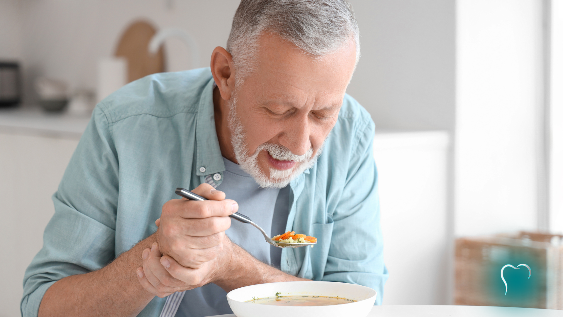 Elderly man in a light blue shirt eating soup at a table, leaning forward with a spoon.
