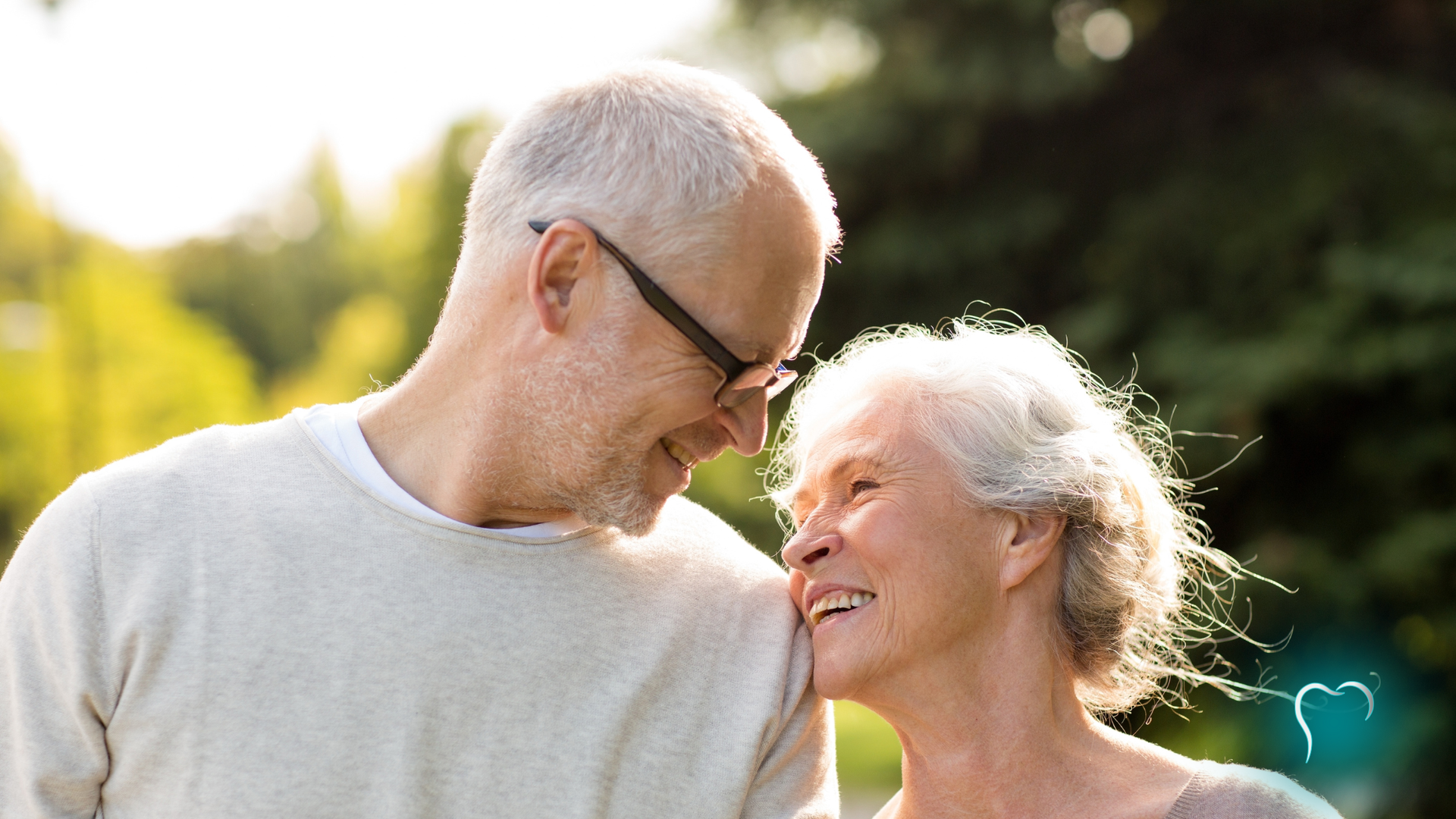 Two smiling people leaning close outdoors in soft sunlight, with greenery in the background.