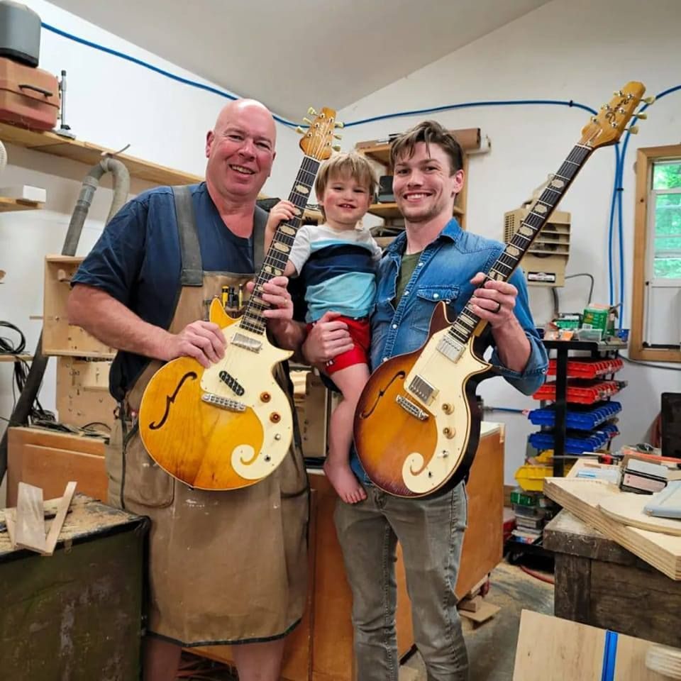Three people in a workshop holding yellow electric guitars and smiling, with tools and shelves in the background