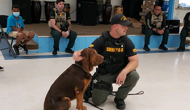 Brown dog sits beside a kneeling handler in a gym, with seated police officers in the background.