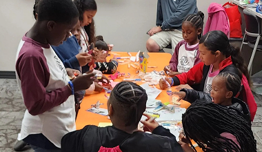 Children sit around a table doing a craft activity with orange paper and supplies.