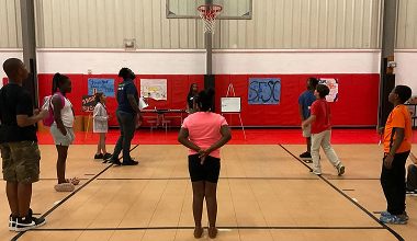 People standing spaced apart in a gym, facing a basketball hoop and red wall during an indoor activity