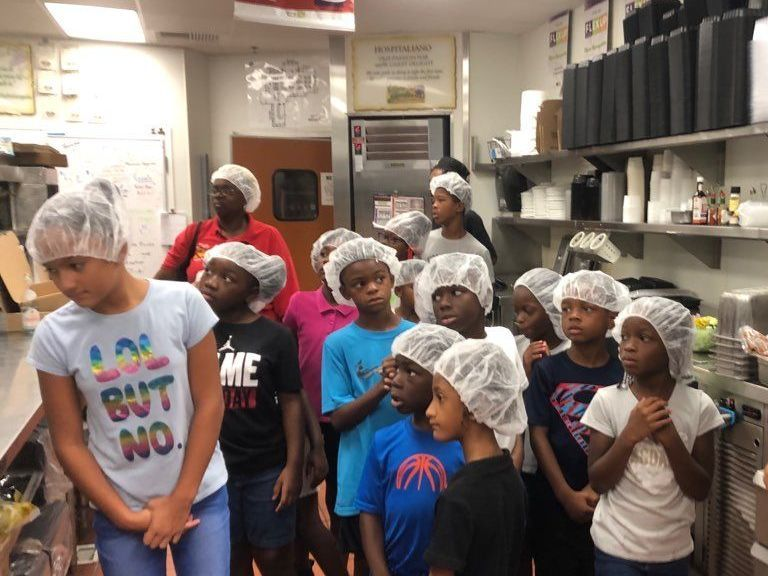 Children in hairnets and aprons standing together in a commercial kitchen, some looking toward the camera.