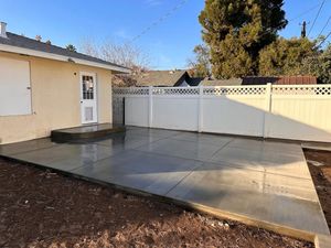 Backyard patio with a concrete slab, white fence, and a beige house under a clear blue sky