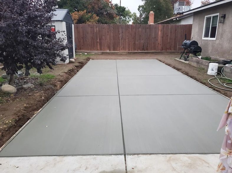 Fresh concrete driveway in a backyard, bordered by dirt and a wooden fence, with a person at the right edge.