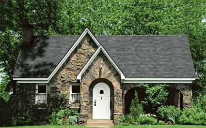 Small stone cottage with white door, dark roof, and lush green trees behind it.