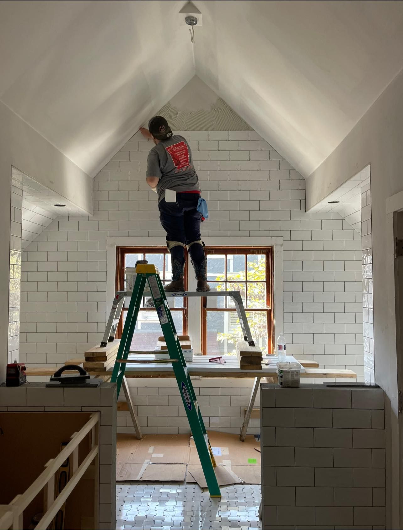 A man is standing on a ladder painting a white brick wall.
