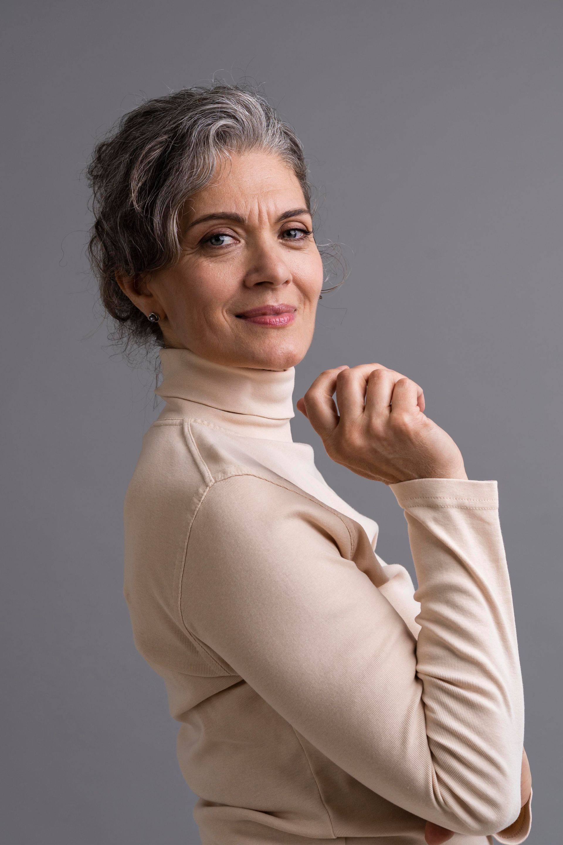 Una mujer de cabello gris con un cuello alto de color canela sonríe, con la mano en la barbilla, sobre un fondo gris.