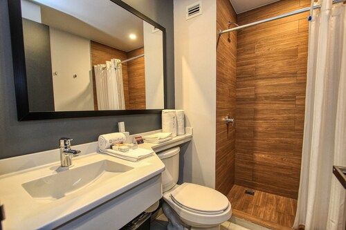 Baño con lavabo, inodoro y ducha blancos con azulejos imitación madera. El espejo refleja la ducha y el lavabo.