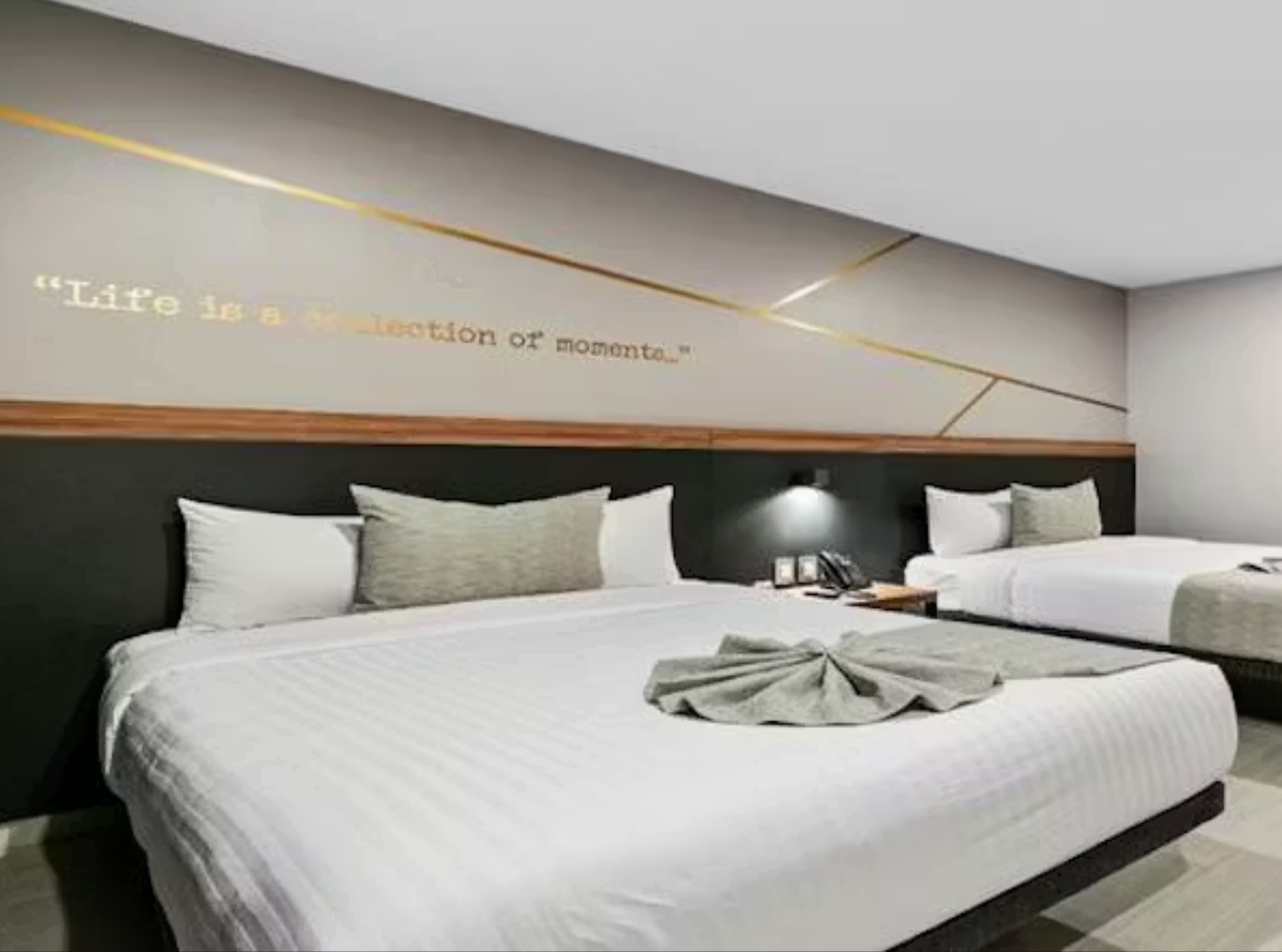 Habitación de hotel con dos camas, ropa de cama gris y blanca, pared decorativa con la cita