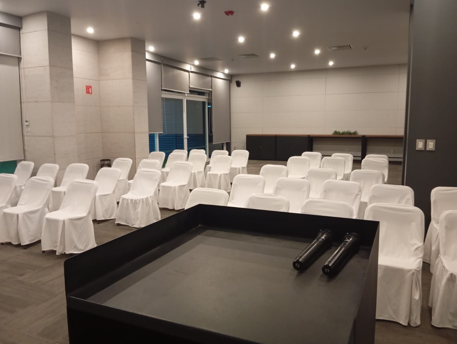 Sala de conferencias vacía con filas de sillas cubiertas de blanco frente a un podio negro con micrófonos.