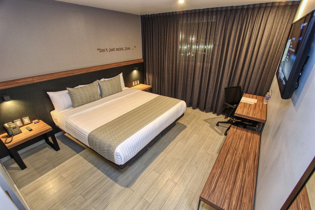Habitación de hotel con cama, escritorio, TV, paredes grises y cortinas oscuras.